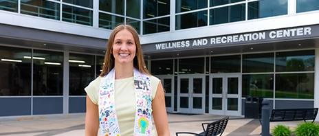 Abby Wolter, '26, smiles into the camera in front of St. Ambrose University's Wellness and Recreation center.