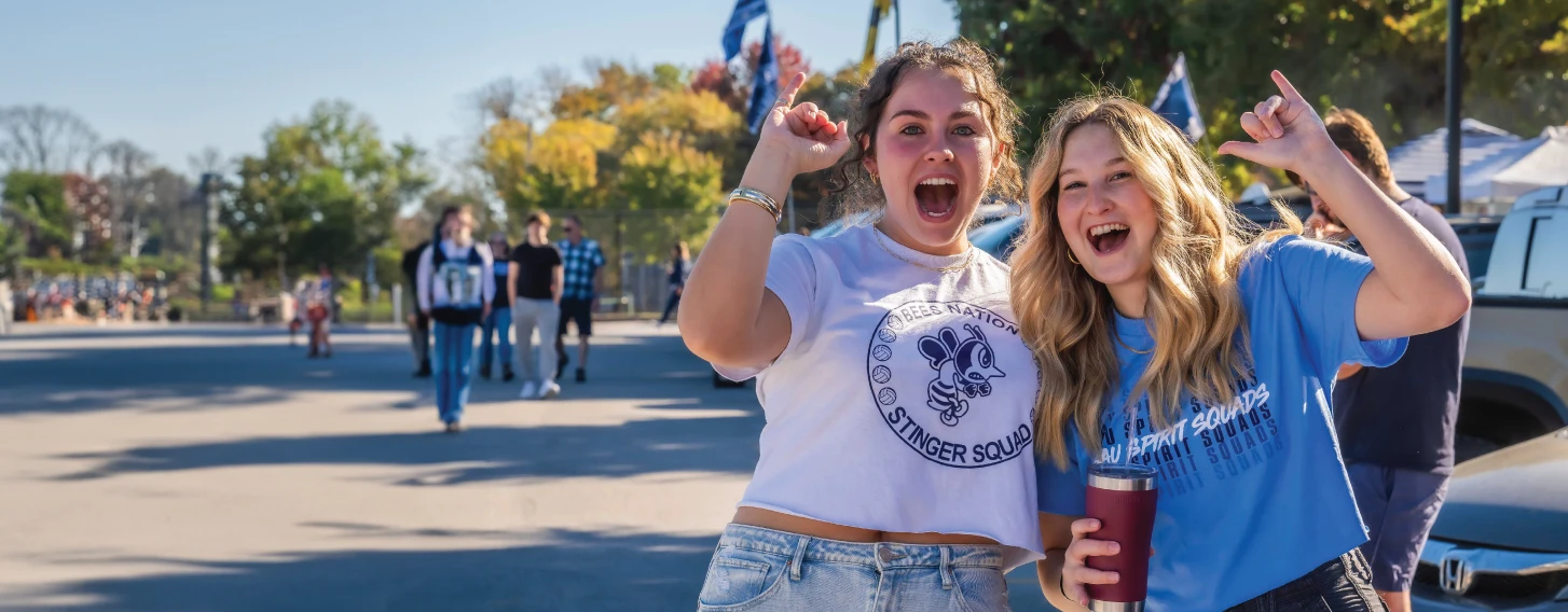 Two smiling students outdoors holding their pinky fingers up in a
